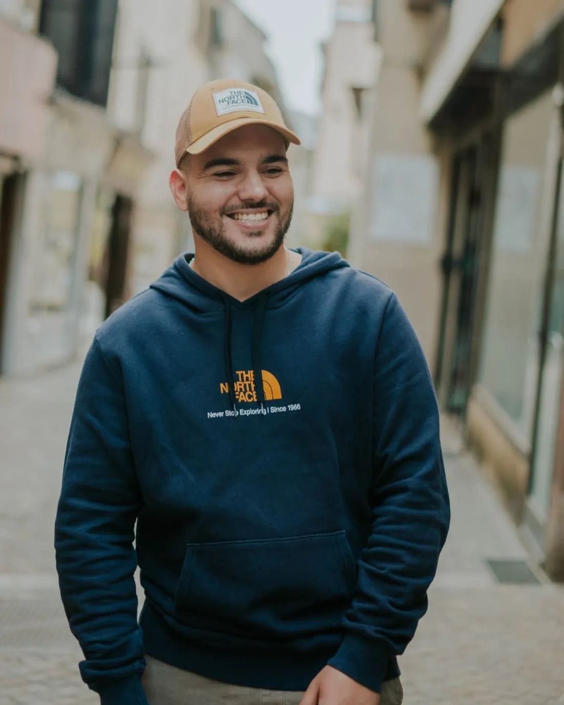 Antoine Bessaou, souriant, en casquette et sweat bleu marine dans une rue piétonne de Rodez